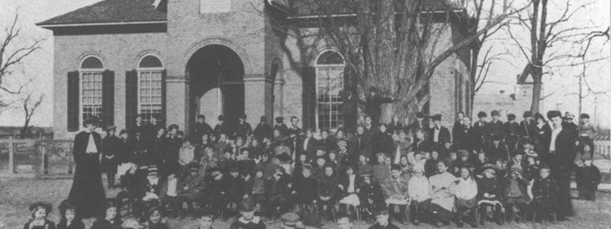 Small brick building in background with a large group of students sitting outside