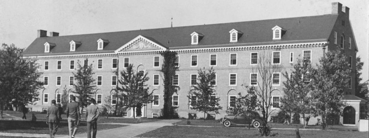 Black and white photo of the four stotry brick dorm with car parked out front and three students walking
