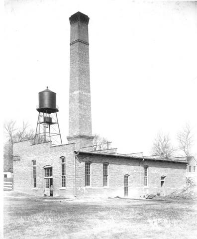 Black and white photo of the brick Power Plant with tall chimney stack and water tower in the distance