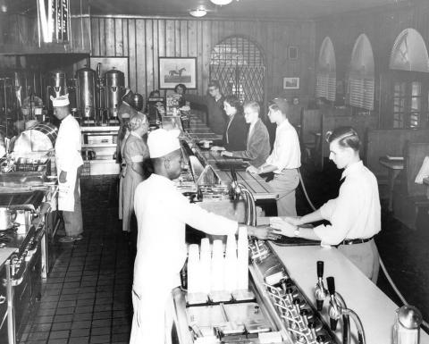 Black and white photo, circa 1950s, with staff preparing meals in the kitchen on the left and students picking up meals cafeteria style on the right