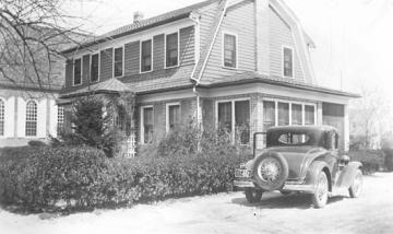 Black and white photo of two story colonial house with 1950s era car parked beside it