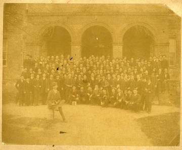 Black and white photo of large group of students sitting on the steps of a building with one sitting in front on a boundary stone