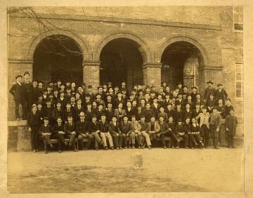 Black and white photo of large group of students posing in front of a building with a boundary stone in front