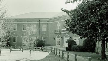 Black and white photo of two story, brick Campus Center with three arched doors and wood fenced sidewalk