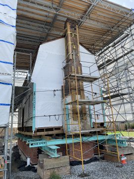 white frame building with brick chimney resting on steel beams and surrounded by scaffolding and plastic sheeting